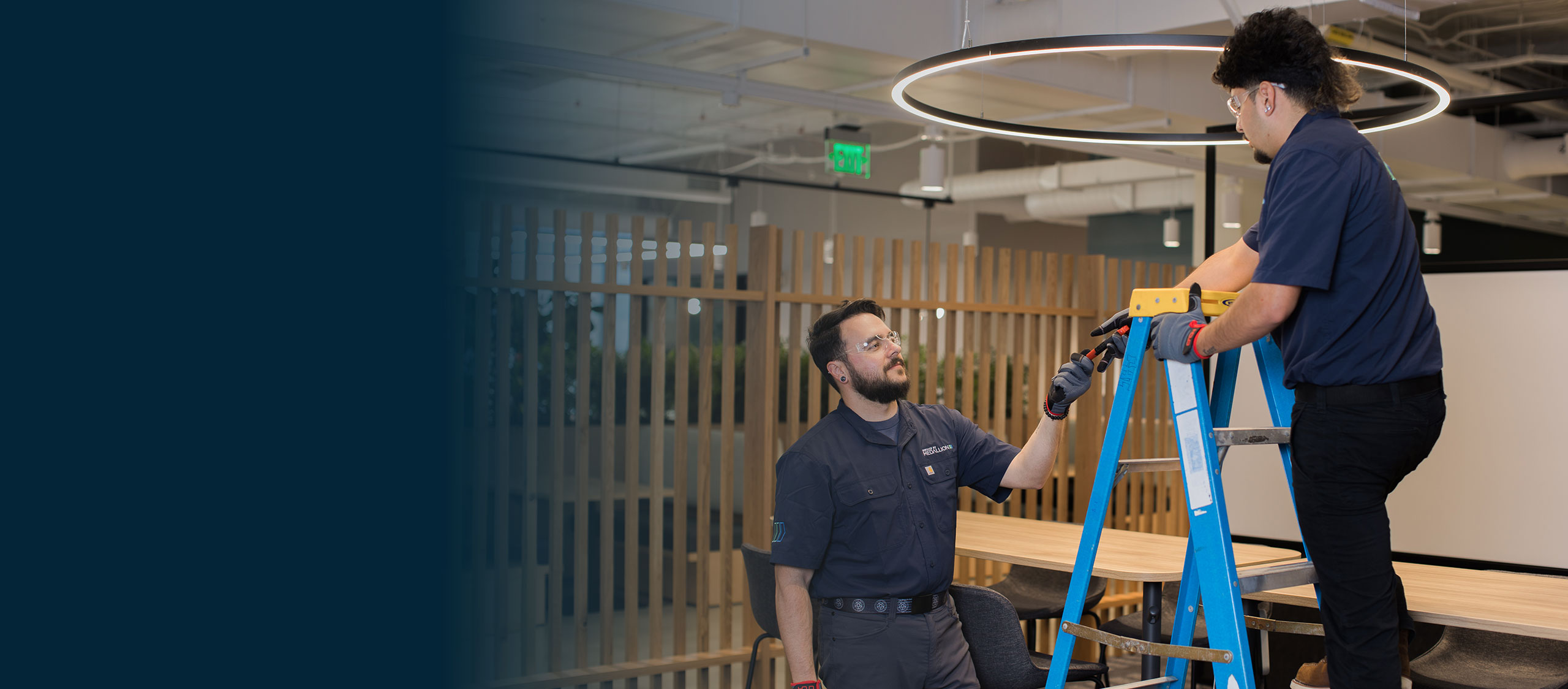 Facility maintenance technicians working together on a ladder to perform building maintenance in a modern office environment