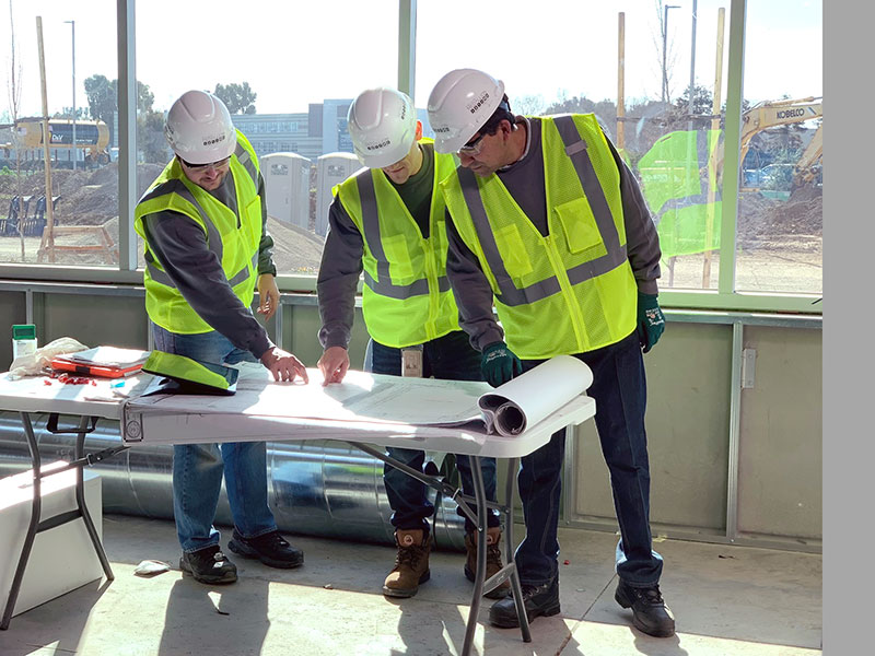 Three construction workers in safety vests and hard hats reviewing plans on a table