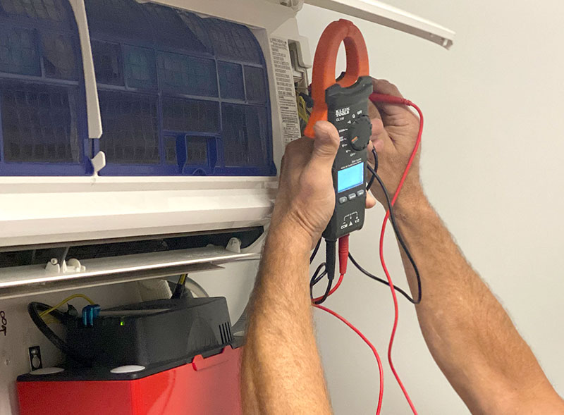 Close-up of an facility technician performing electrical diagnostics on a wall-mounted mini-split air conditioning system using a digital clamp meter