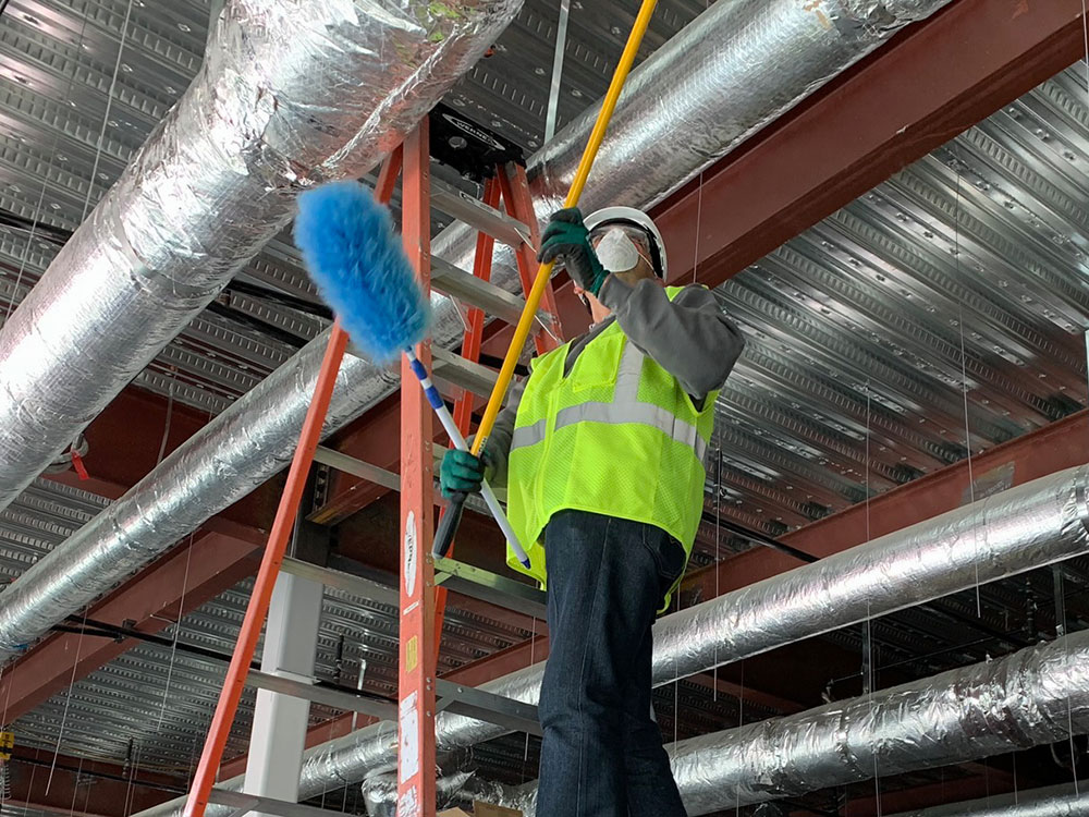 Facility services technician performing high dusting and overhead cleaning around exposed ductwork and ceiling infrastructure