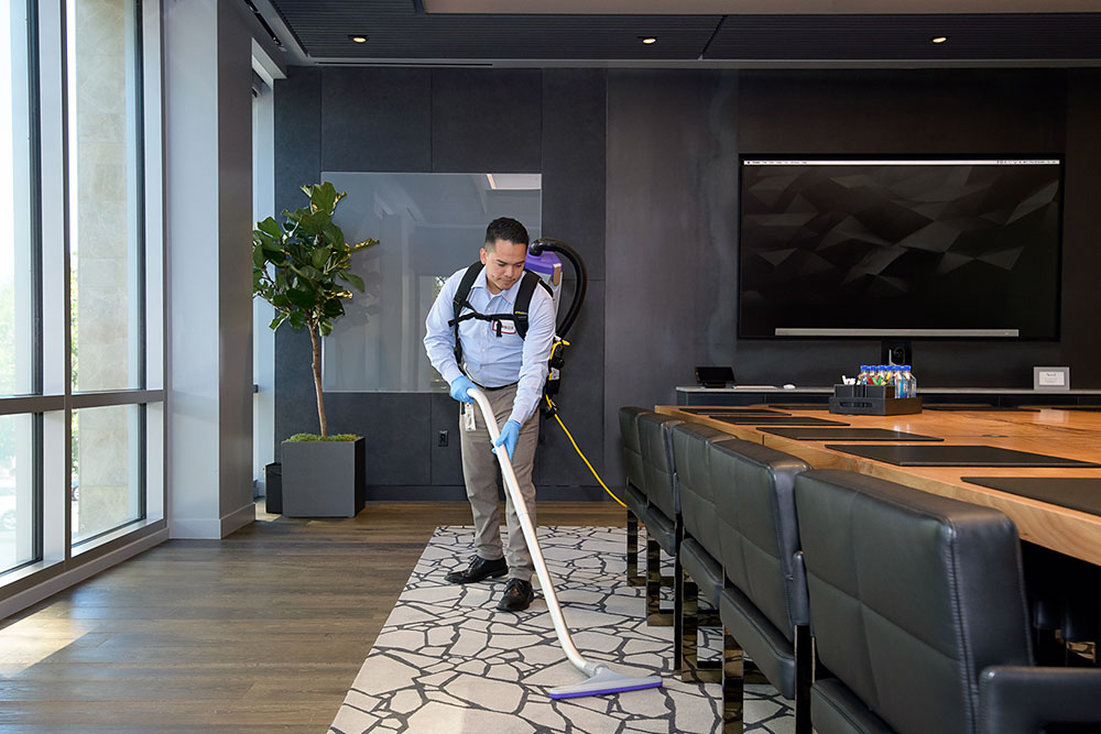 Janitor performing floor care with a backpack vacuum in an office conference room
