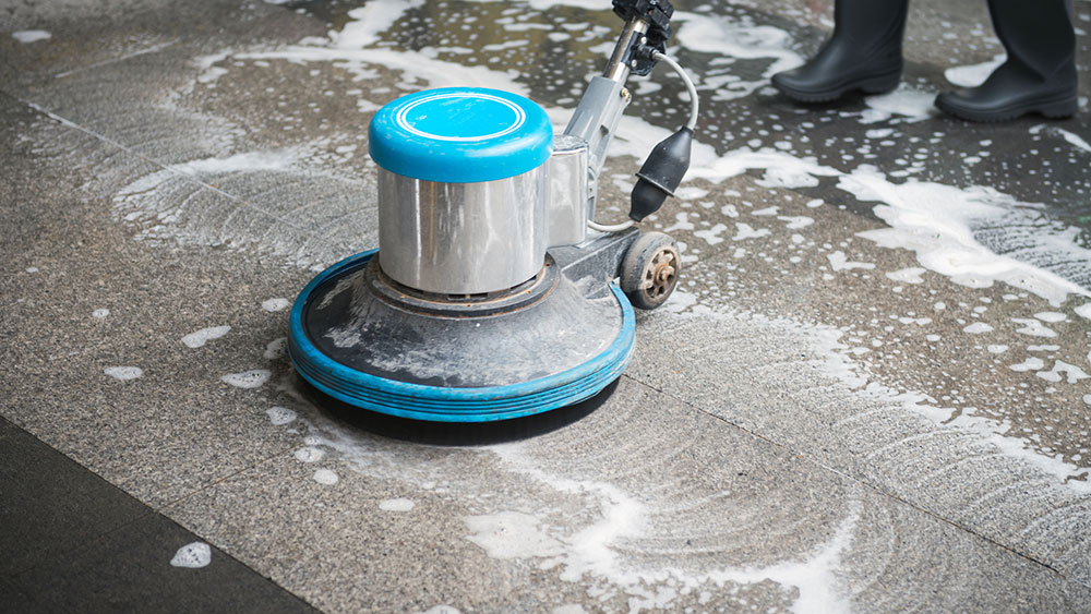 Commercial floor cleaning machine scrubbing a hard surface floor with cleaning solution during floor maintenance.