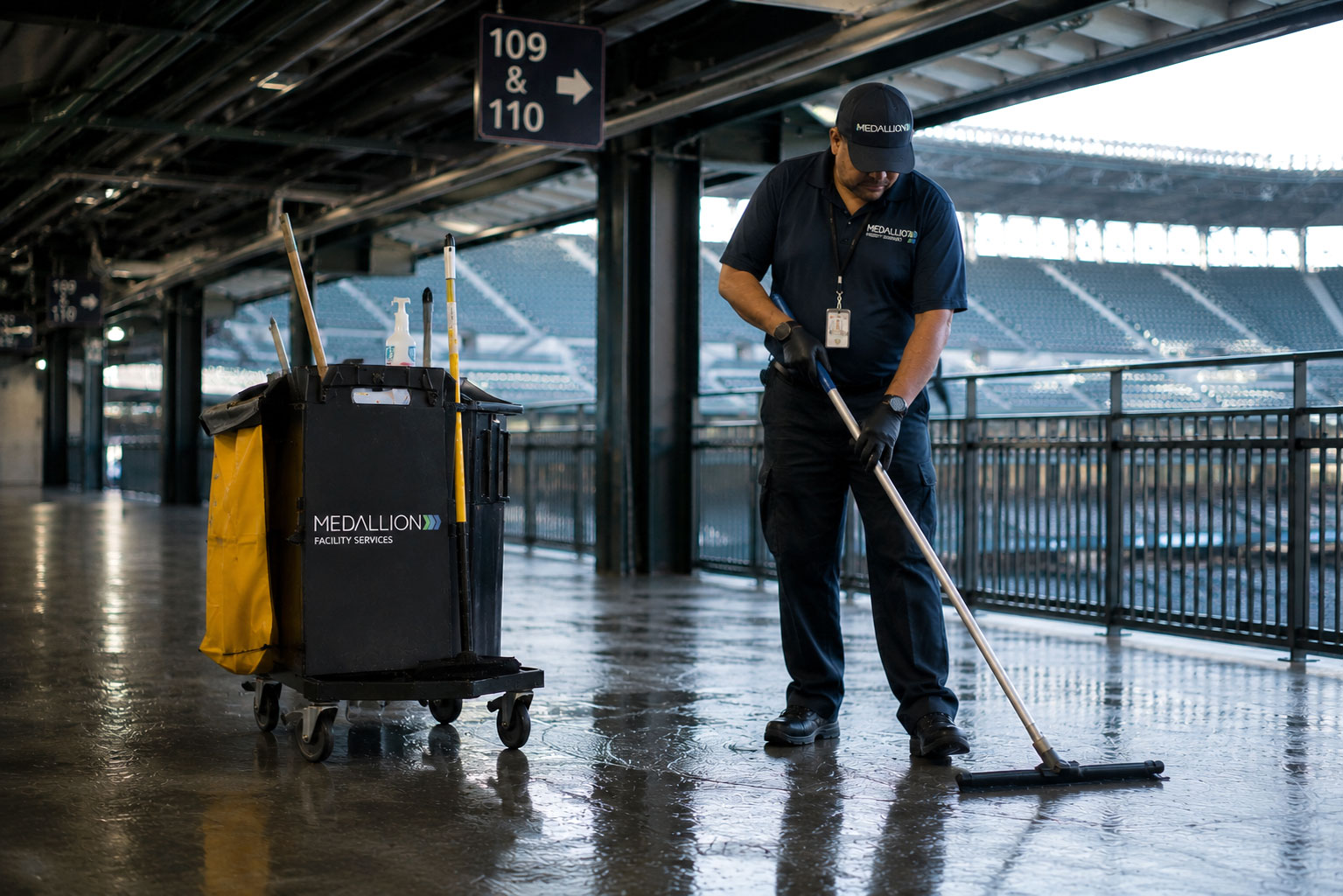 Janitorial professional cleaning a stadium concourse floor with a floor squeegee during facility maintenance operations
